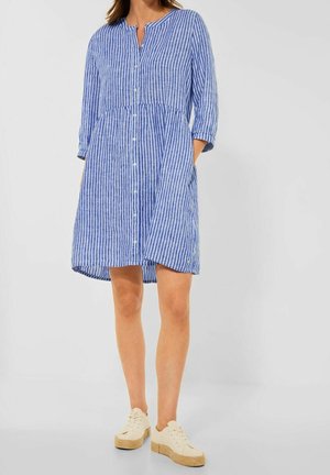 Mujer con un vestido abotonado de rayas verticales azules y blancas, con mangas tres cuartos y zapatillas blancas, de pie con las manos en los bolsillos.