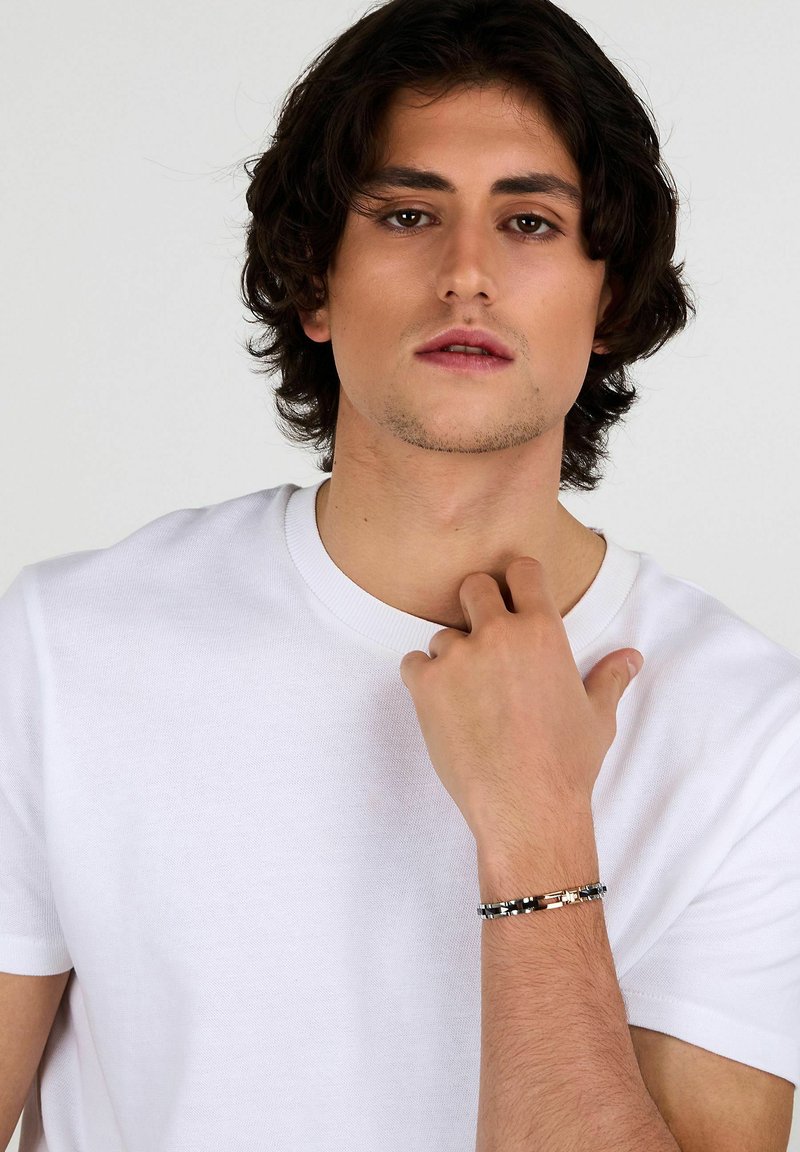 Bracciale argento e nero con intricati anelli di metallo, indossato al polso. Accompagnato da una maglietta bianca semplice con scollo rotondo.