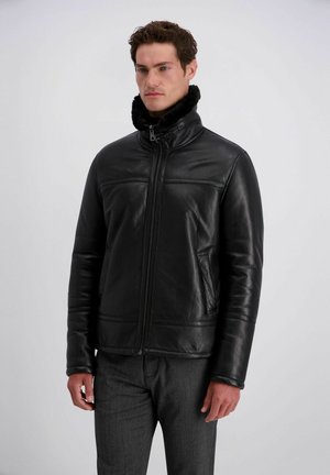 Mann trägt eine schwarze Lederjacke mit Fellkragen und graue Hose, steht vor einem schlichten hellen Hintergrund.