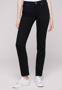 Jeans negros ajustados con cinco bolsillos, cierre de cremallera y botón. La tela tiene una textura suave. Se combinan con zapatillas blancas.