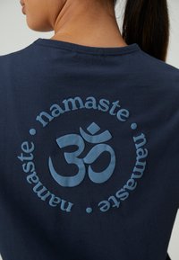 Tamsiai mėlynos spalvos medvilninė marškinėliai su apvalia pakelta grafika, kurioje pavaizduotas "Om" simbolis ir žodis "namaste" šviesiai mėlyna spalva.