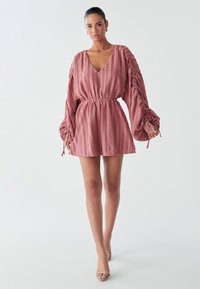 Model trägt ein pinkfarbenes, strukturiertes Minikleid mit Puffärmeln und tailliertem Bund, kombiniert mit Riemchenheels, und steht vor einem hellen Hintergrund.