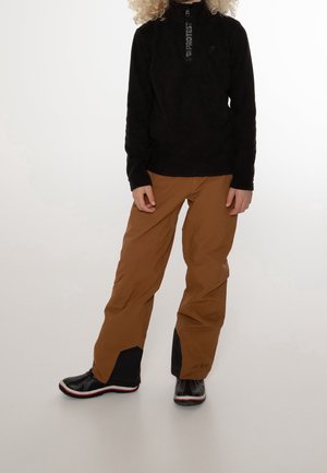 Personne portant un pull-over noir à manches longues avec une fermeture éclair et un pantalon marron rentré dans des bottes d'hiver noires à semelles rouges, debout devant un fond uni.