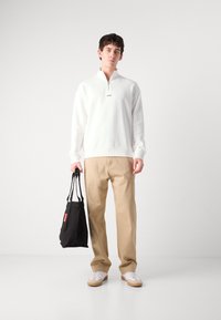 Hvit zip-up hettegenser med logo, beige bukser og en svart totebag med rød tag, kombinert med hvite sneakers. Enkelt, avslappet antrekk.