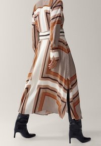 Robe longue à motifs présentant des rayures géométriques marron, orange et blanche. Le tissu est lisse ; le design comporte une fente latérale et une ceinture.