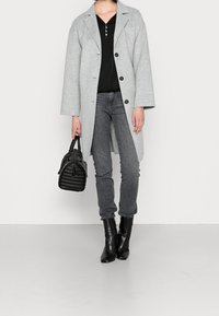 Persona che indossa un cappotto grigio chiaro con bottoni sopra una maglia nera, jeans grigi e stivali neri alla caviglia, mentre tiene una borsa nera con manici superiori.