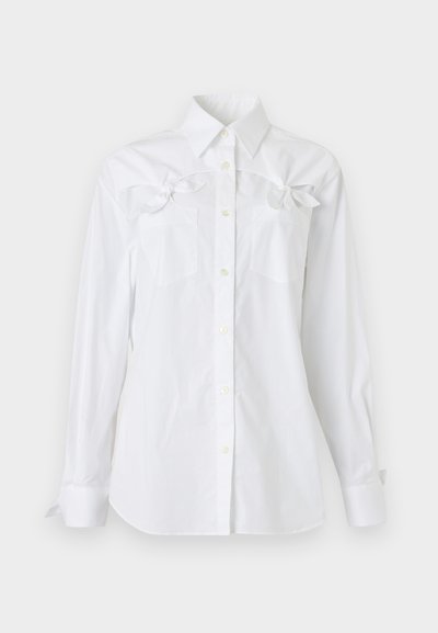 Chemise blanche à manches longues, boutonnée, avec un col et deux poches poitrine, chacune ornée d'un noeud décoratif près du haut.