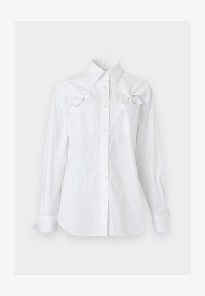 Chemise blanche à manches longues, boutonnée, avec un col et deux poches poitrine, chacune ornée d'un noeud décoratif près du haut.