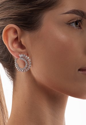 Strieborná náušnica v tvare kruhu s vírivým dizajnom, zdobená malými priehľadnými kryštálmi a hruškovými akcentmi, na neutrálnom odtieni pleti.