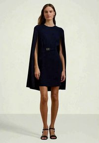 Γυναίκα που στέκεται φορώντας ένα μαύρο φόρεμα με κάπα και ζώνη στη μέση, καθώς και μαύρες ανοιχτές παντόφλες, σε έναν απλό φωτεινό φόντο.