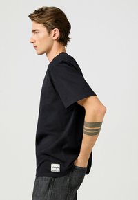 Sort bomulds T-shirt med korte ærmer, der har en rund hals og et rektangulært Wrangler-logo label nær kanten. Båret med grå jeans.