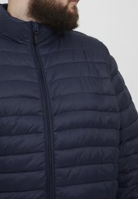 Mornarska pufasta jakna s horizontalnim kvadratnim dizajnom, visokim ovratnikom i crnim patentnim zatvaračem. Glatka tekstura; lagan i izoliran izgled.