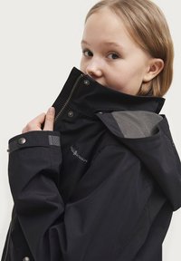 Zwarte waterdichte jas met hoge kraag, zilveren rits en gaasaccenten op de schouders. Beschikt over drukknoopsluitingen en een logo op de mouw.