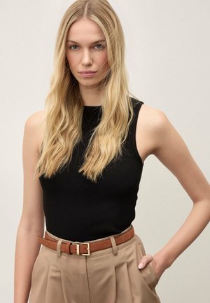 Mujer rubia con cabello largo que lleva una camiseta negra sin mangas metida en unos pantalones beige de talle alto con un cinturón marrón, con la mano en el bolsillo.