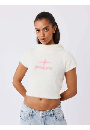Jeune femme aux longs cheveux foncés porte un top court blanc duveteux avec un logo rose et un jean en denim bleu clair, posant les mains derrière le dos.