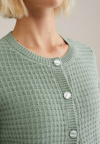Cardigan en tricot vert menthe avec un motif texturé, col rond et grands boutons clairs sur le devant.