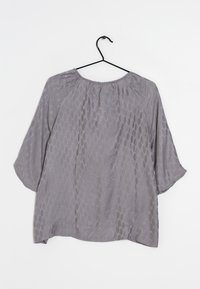 Šedá, texturovaná blůza s hexagonálním vzorem, lehký materiál, volný střih a široké tříčtvrteční rukávy. Visí na černém háčku.
