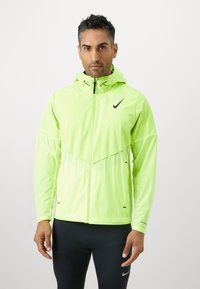 Giacca leggera di colore verde neon con cappuccio, dotata di chiusura con zip e logo Nike nero. Tessuto testurizzato con vestibilità atletica e dettagli riflettenti.