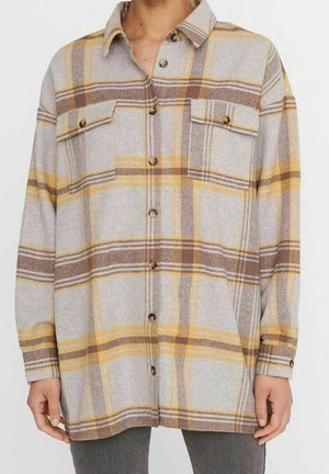 Koszula w kratę na guziki z długim rękawem i kieszeniami na piersi, z beżowymi, brązowymi i żółtymi paskami, noszona na ciemnych spodniach.