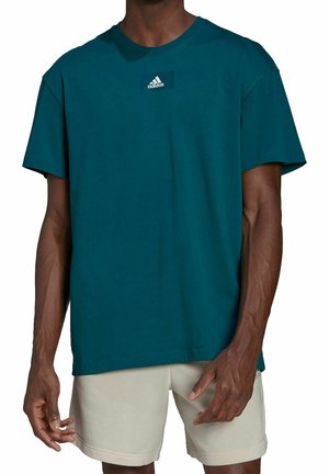 Muž v tyrkysovém tričku Adidas s logem na hrudi a béžových kraťasech, stojící s rukama volně podél těla.