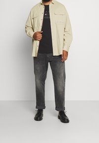 Jasnobeżowa koszula na guziki z dwoma kieszeniami na piersi, noszona na czarnym t-shircie z nadrukiem. Ciemnoszare zwężane jeansy i czarne buty na sznurowadła.