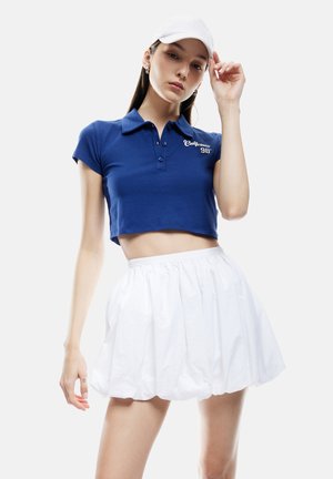 Kobieta w białej czapce, ubrana w niebieskie krótkie polo z napisem "California 98" oraz białą plisowaną spódnicę z wysokim stanem, pozująca na jednolitym tle.