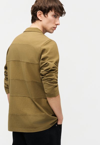 Νεαρός με καστανά μαλλιά που φοράει πουκάμισο μακρυμάνικο σε ελιά πράσινη απόχρωση, στέκεται πλαγίως με το ένα χέρι στην τσέπη, μπροστά σε ένα απλό φόντο.