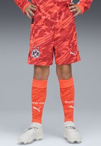 Shorts de sport orange vif avec un motif tourbillonnant et le logo BVB, associés à des chaussettes montantes assorties et des crampons de football de couleur claire.