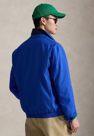 Μπλε bomber μπουφάν με απαλό ύφασμα, ελαστική μέση και γιακά. Φορεμένο με πράσινο καπέλο και γυαλιά ηλίου, παρουσιάζοντας μια casual σιλουέτα.