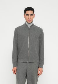 Uomo che indossa una giacca grigia con zip su un dolcevita grigio chiaro, con pantaloni grigi abbinati, in piedi contro uno sfondo bianco.