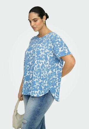 Kvinde med mørkt hår sat op i en knold, iført en blå og hvid blomstret kortærmet top og lyseblå slidte jeans, der holder en beige håndtaske.