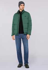 Zelená péřová bunda s vysokým límcem, spárovaná s černou zipovou mikinou, modrými džíny a černými botami. Model nosí šedou čepici.