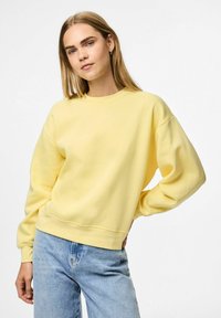 Felpa gialla con collo rotondo e maniche lunghe e larghe. Materiale in cotone morbido con una superficie liscia. Indossata con jeans azzurri chiari.