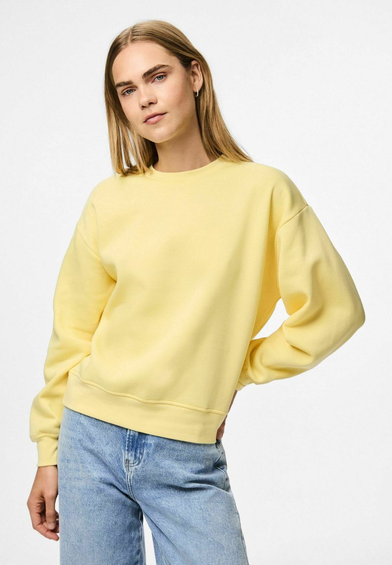 Felpa gialla con collo rotondo e maniche lunghe e larghe. Materiale in cotone morbido con una superficie liscia. Indossata con jeans azzurri chiari.