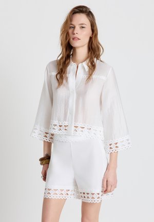 Frau mit langem, welligem Haar, die eine weiße Bluse mit Knöpfen und Shorts mit Spitzenbesatz trägt, steht vor einem schlichten hellen Hintergrund.