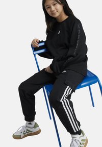 Sudadera negra con logo de Adidas, pantalones cargo negros con rayas blancas y zapatillas verdes y blancas. Sujeto sentado en una silla azul.