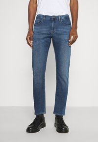 Man som bär slim-fit blå jeans med subtila blekningar, svarta sneakers, står mot en enfärgad vit bakgrund.