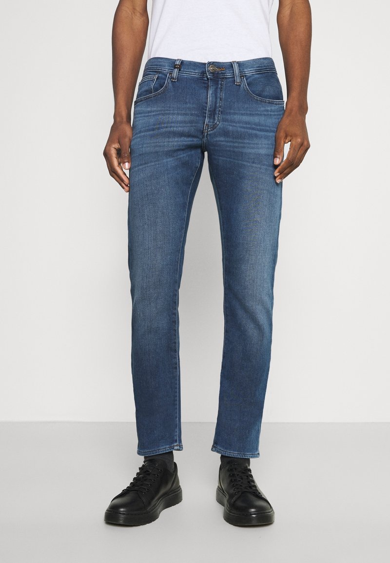 Man som bär slim-fit blå jeans med subtila blekningar, svarta sneakers, står mot en enfärgad vit bakgrund.