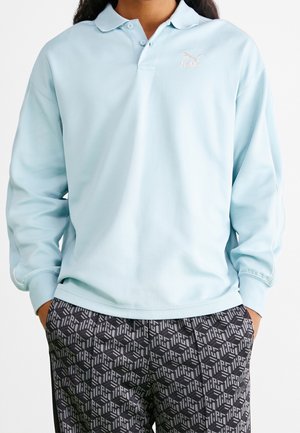 Person iført en lyseblå langærmet polo og sort mønstrede bukser med hænderne i lommerne foran en hvid baggrund.