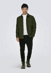 Joven con un gorro negro, camiseta blanca, chaqueta verde oliva, pantalones negros y botas verde oliva, de pie con una mano en el bolsillo.