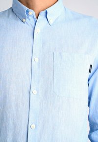 Camisa de botones azul claro hecha de tela texturizada, con un bolsillo en el pecho y botones blancos en el frente.