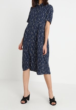 Kobieta w granatowej sukience midi zapinanej na guziki z białymi pionowymi wzorami w paski i czarnych sandałach na obcasie z odkrytymi palcami.