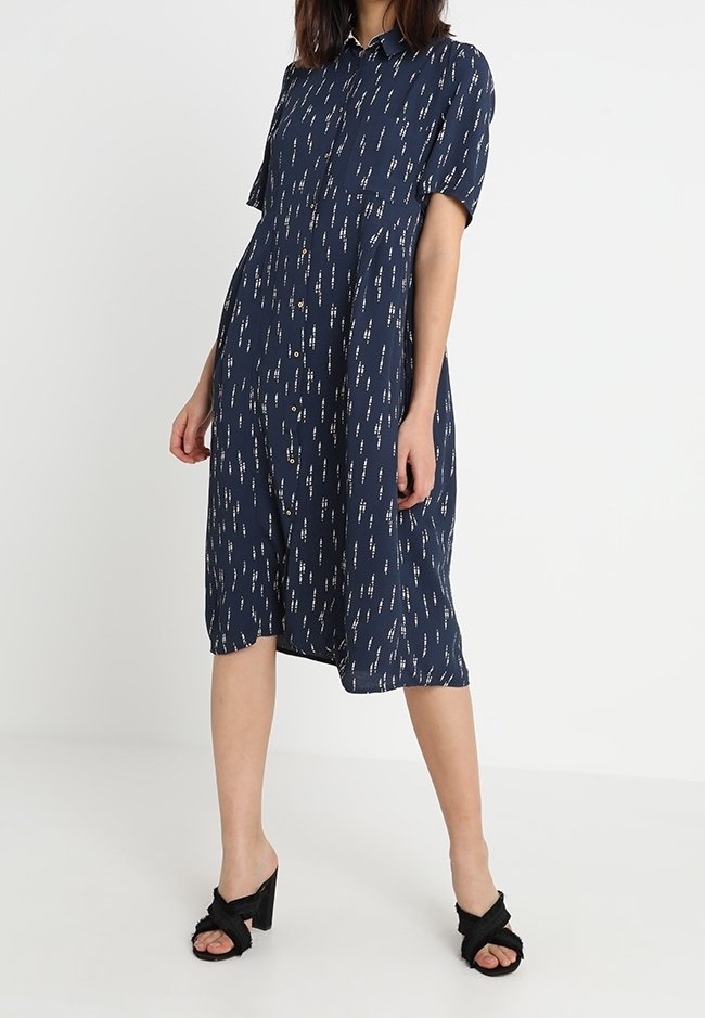 Femme portant une robe midi bleu marine boutonnée avec des motifs de tirets verticaux blancs et des sandales noires à talons ouvertes.