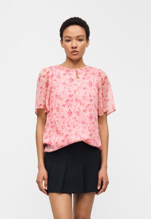 Kobieta ubrana w różową, kwiatową bluzkę z krótkim rękawem i czarną plisowaną mini spódnicę, stojąca na tle jednolitego, jasnoszarego tła.