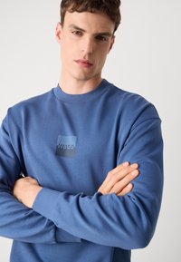 Modrá mikina vyrobená z hladkého materiálu, s obdĺžnikovou nášivkou loga s nápisom "HUGO." Dizajn obsahuje rebrované manžety a výstrih.