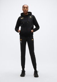 Sweat à capuche noir avec un design texturé et des accents jaunes, associé à un pantalon noir avec des éléments de marque. Confectionné en tissu doux, idéal pour une tenue décontractée.