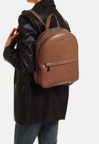 Zaino in pelle marrone dalla forma arrotondata, dotato di doppia cerniera, tasca frontale e spallacci regolabili, indossato sopra una giacca di pelle nera.