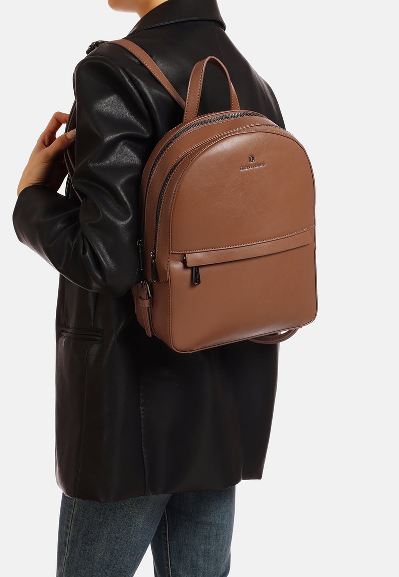 Zaino in pelle marrone dalla forma arrotondata, dotato di doppia cerniera, tasca frontale e spallacci regolabili, indossato sopra una giacca di pelle nera.