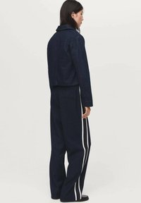 Persoon met lang, donker haar, gekleed in een donkerblauwe jas en wijde marineblauwe broek met witte zijsstripes, staande tegen een effen lichte achtergrond.