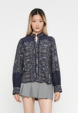 Kvinne med langt hår iført en mørk blomstrete langermet bluse og grå plisserte miniskjørt, stående mot en ensfarget hvit bakgrunn.